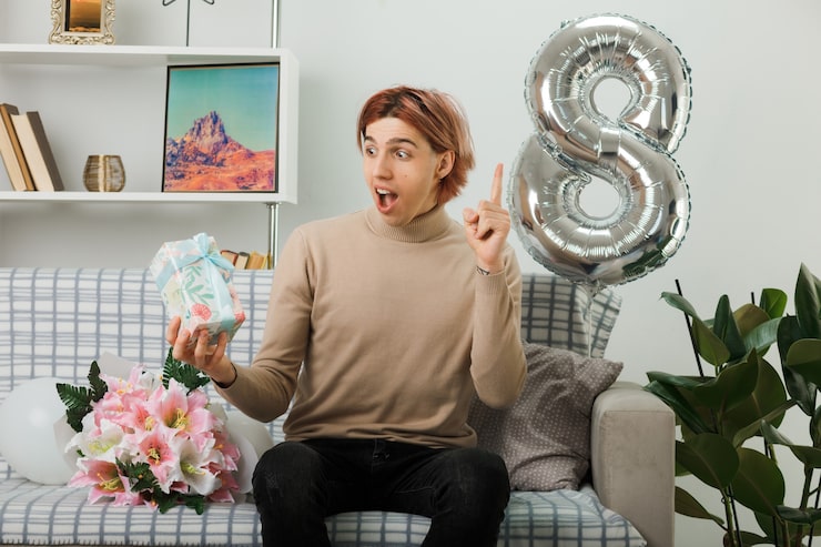 excited-points-up-handsome-guy-happy-women-day-holding-present-sitting-sofa-living-room_141793-123498